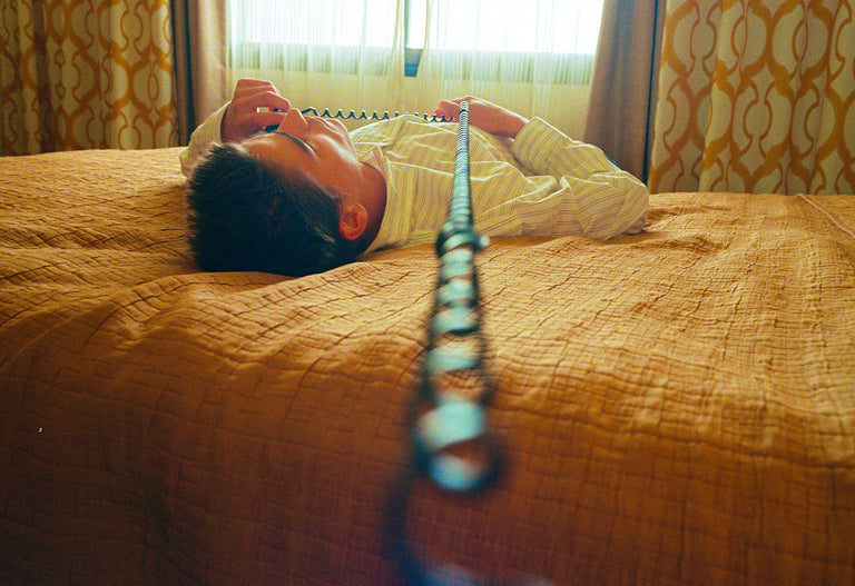 Person lying on a bed with a phone receiver, in a room with patterned curtains.