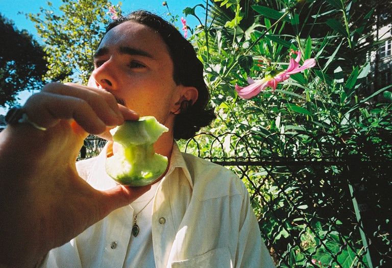 Person holding a green apple outdoors with trees and plants in the background