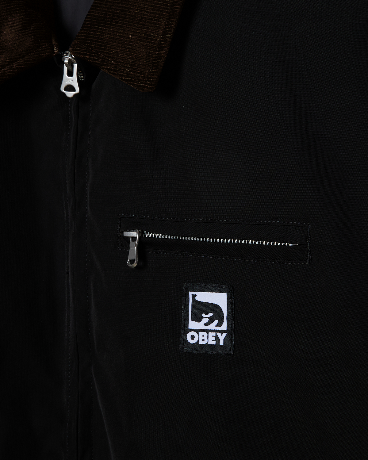 Close-up of a black zip-front jacket with a brown corduroy collar, a zippered chest pocket, and a black-and-white Obey logo patch on the front.