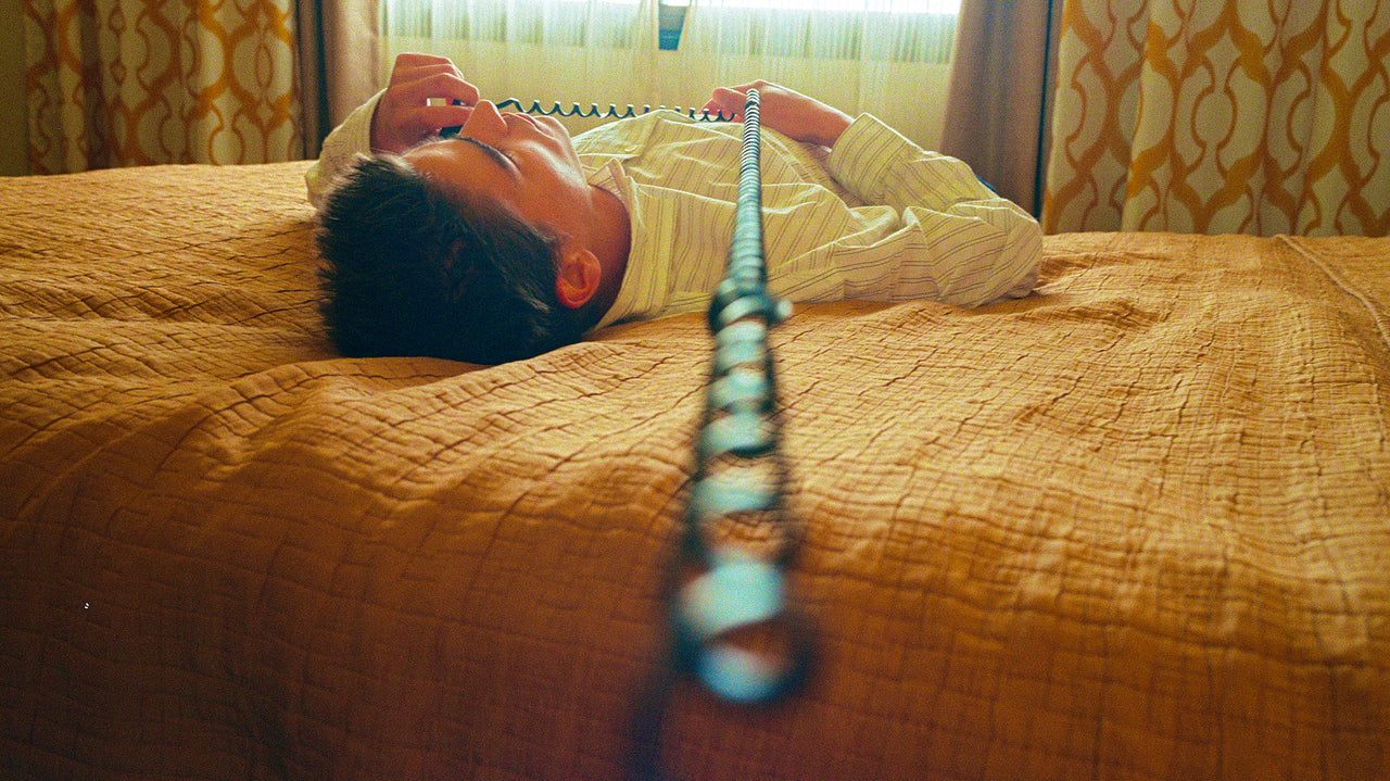 Person lying on a bed with a phone receiver, in a room with patterned curtains.