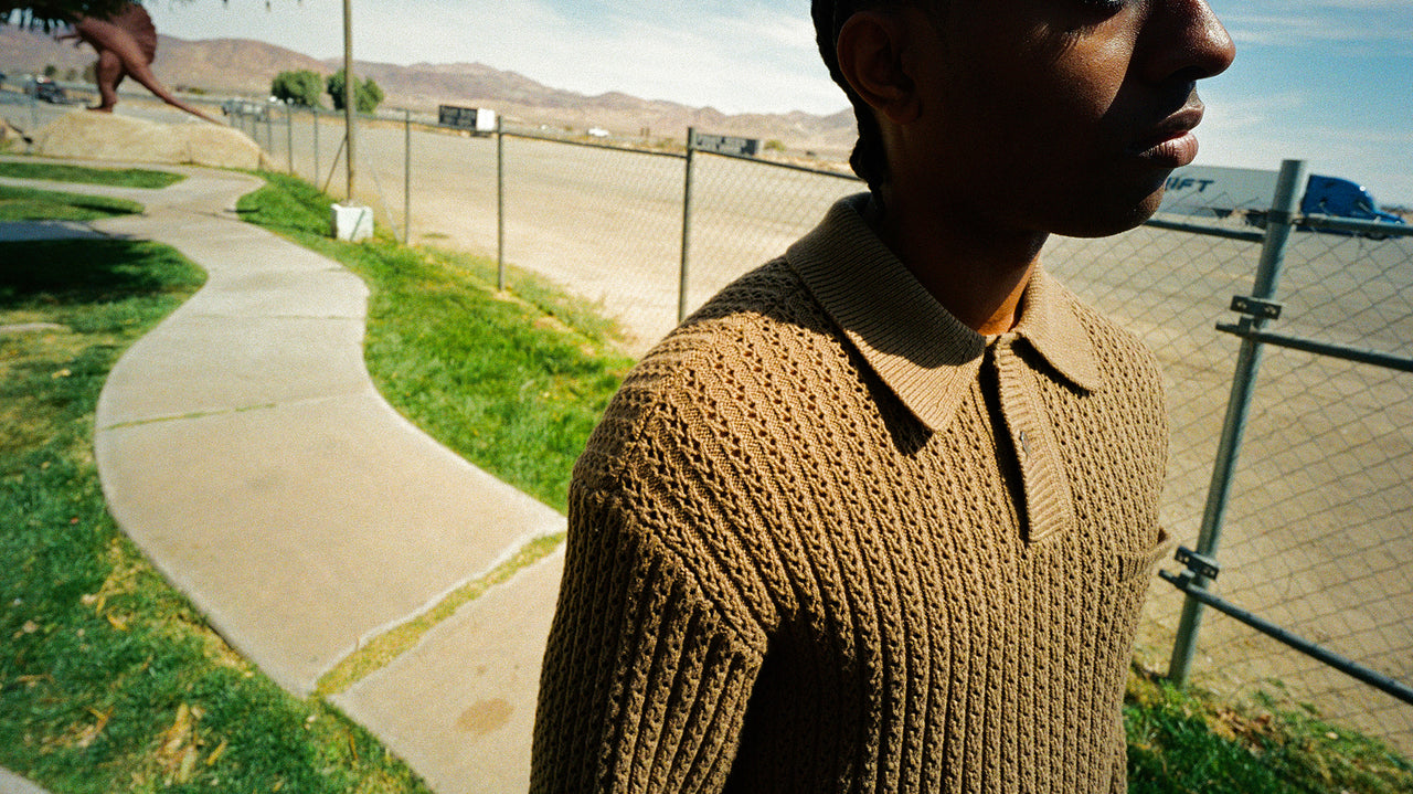 Person wearing a brown knitted sweater standing on a path with grass and a fence in the background.