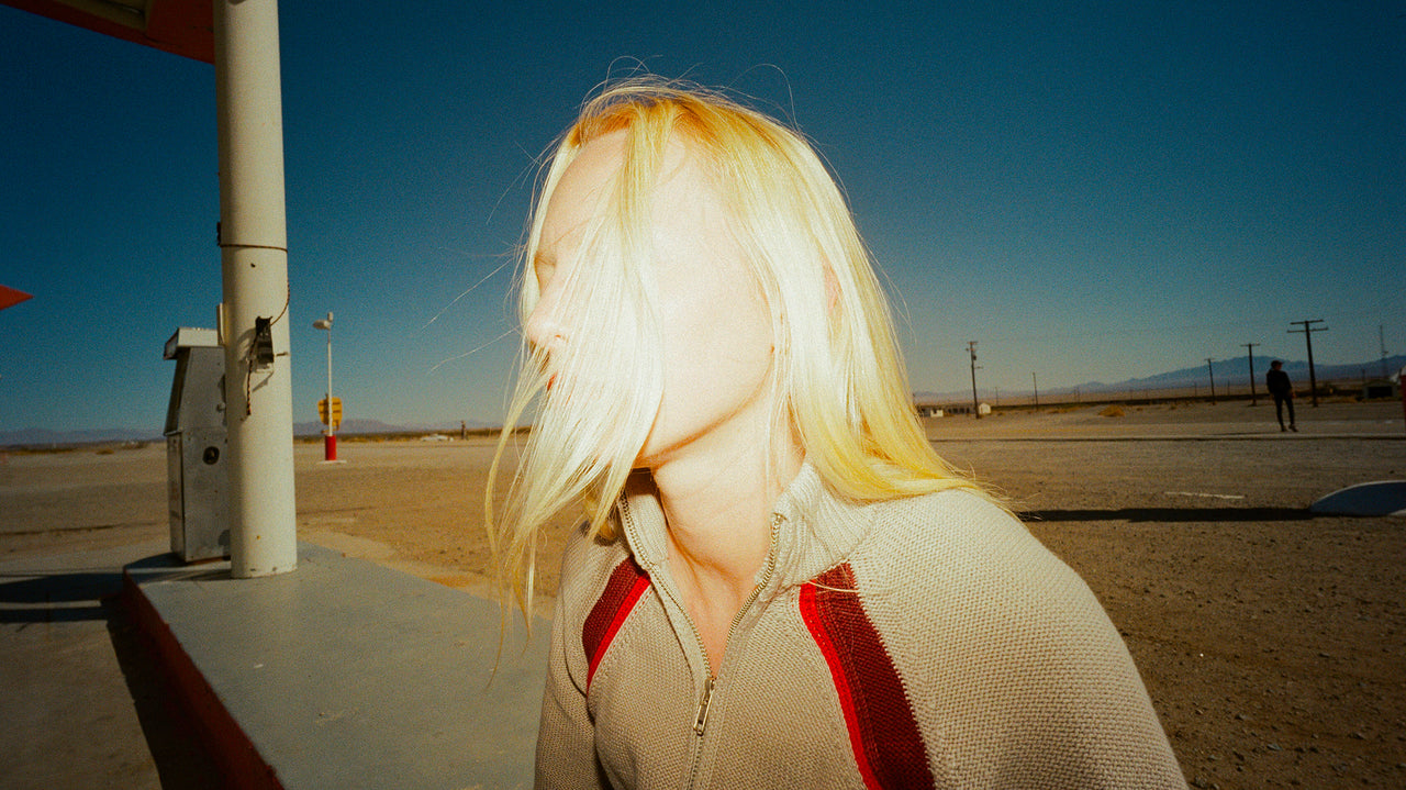 Person with blonde hair standing in a desert-like setting with a clear blue sky.
