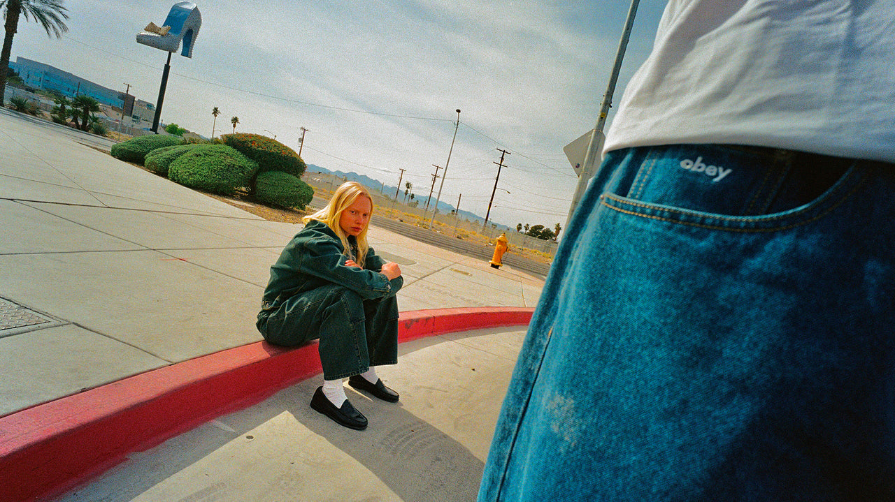 Person sitting on a red curb with a view of a street and buildings in the background.