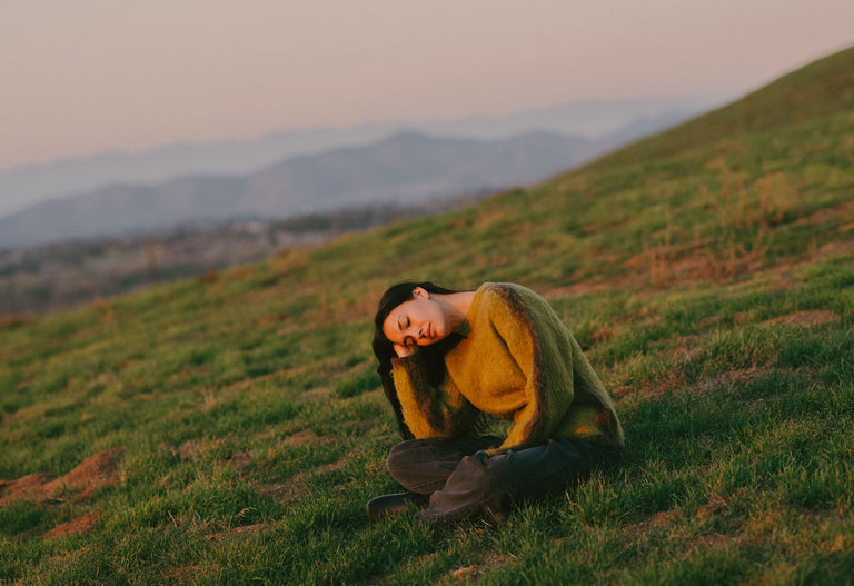 Person sitting on grass wearing a green sweater and brown pants