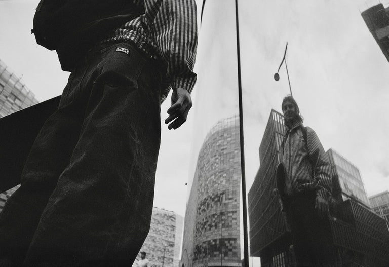 Black and white photo of a person walking on a city street with modern buildings in the background.