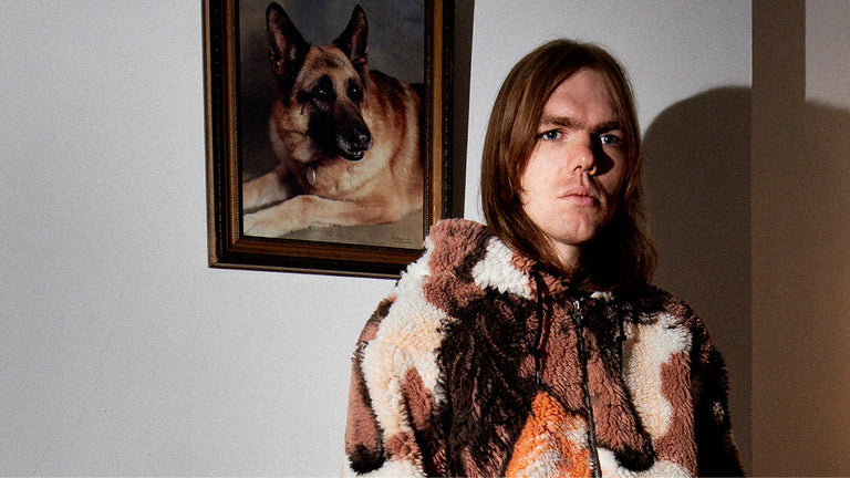 Person wearing a patterned coat standing in front of a wall with a framed picture of a dog.