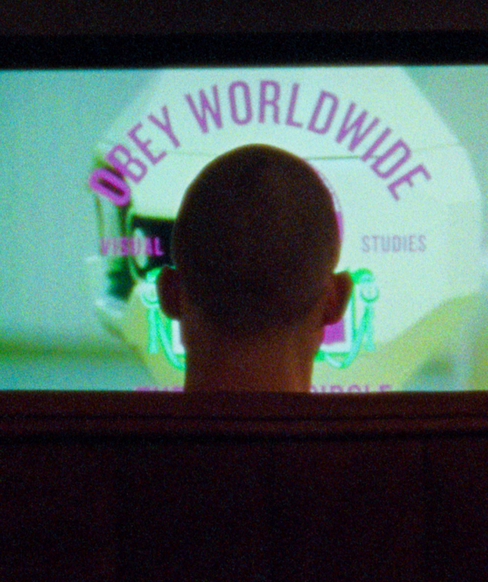 A person viewed from behind, seated in a dark room facing a large screen displaying the text 'OBEY WORLDWIDE' in purple over a pale green graphic.