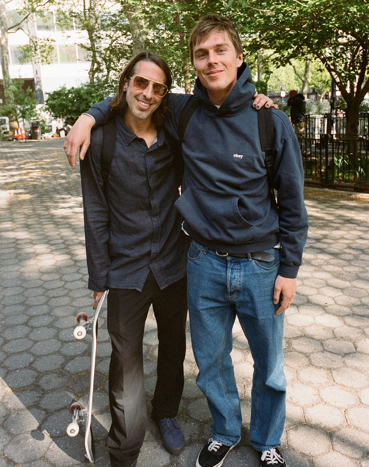 Two men posing together on a sidewalk with a skateboard.