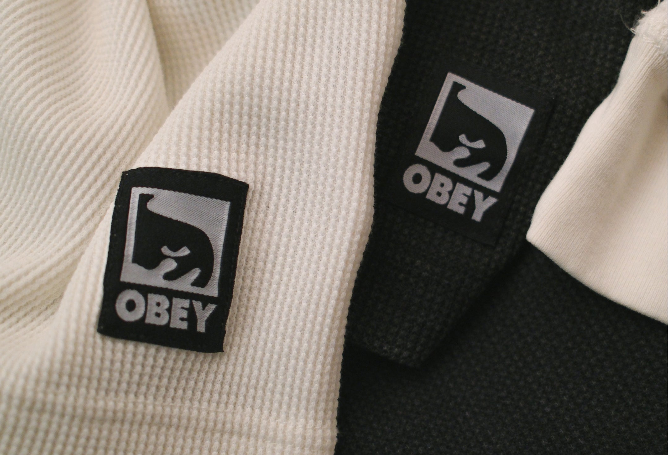 Close-up of a black and white Obey brand garment, showing two square woven logo patches on ribbed fabric.