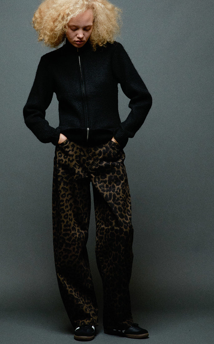 A model with curly blonde hair wearing a black zip-up jacket and leopard-print trousers, standing against a dark gray studio background.