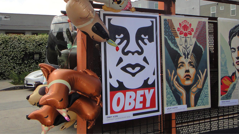 Posters with artistic designs and 'OBEY' branding on a fence, with inflatable dog and cat balloons 