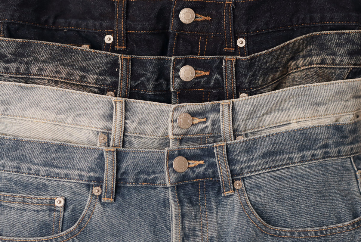 Three pairs of folded denim jeans in different washes stacked together: a dark navy pair at the top, a medium blue pair in the middle, and a light faded blue pair at the bottom. The close-up shows the waistbands, metal buttons, and front pockets.