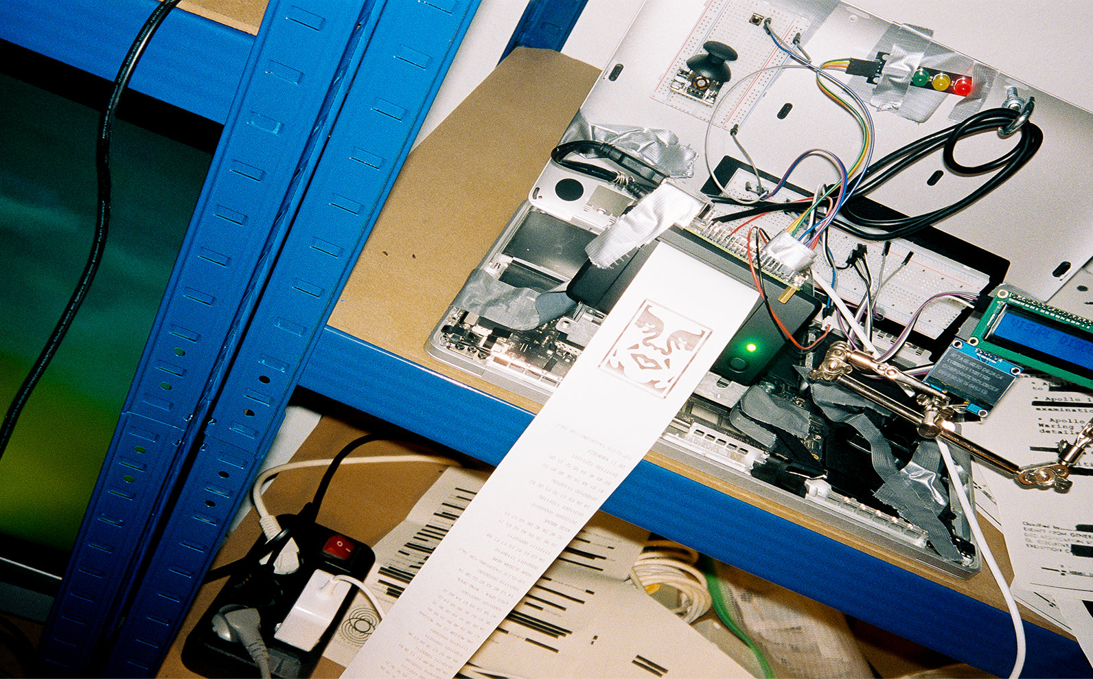 A close-up of an electronic circuit board or DIY electronics project mounted on a blue metal shelf, with numerous wires, components, and a long printed paper trail hanging down with the icon face.