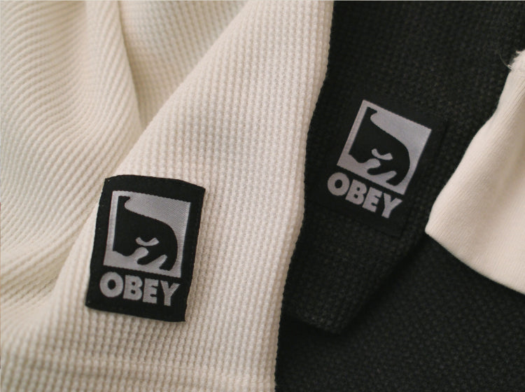 Close-up of a waffle-knit martial arts gi showing two color options: a cream-colored jacket with a black logo patch and a black jacket with a white logo patch. The patch features the Obey brand logo and name.