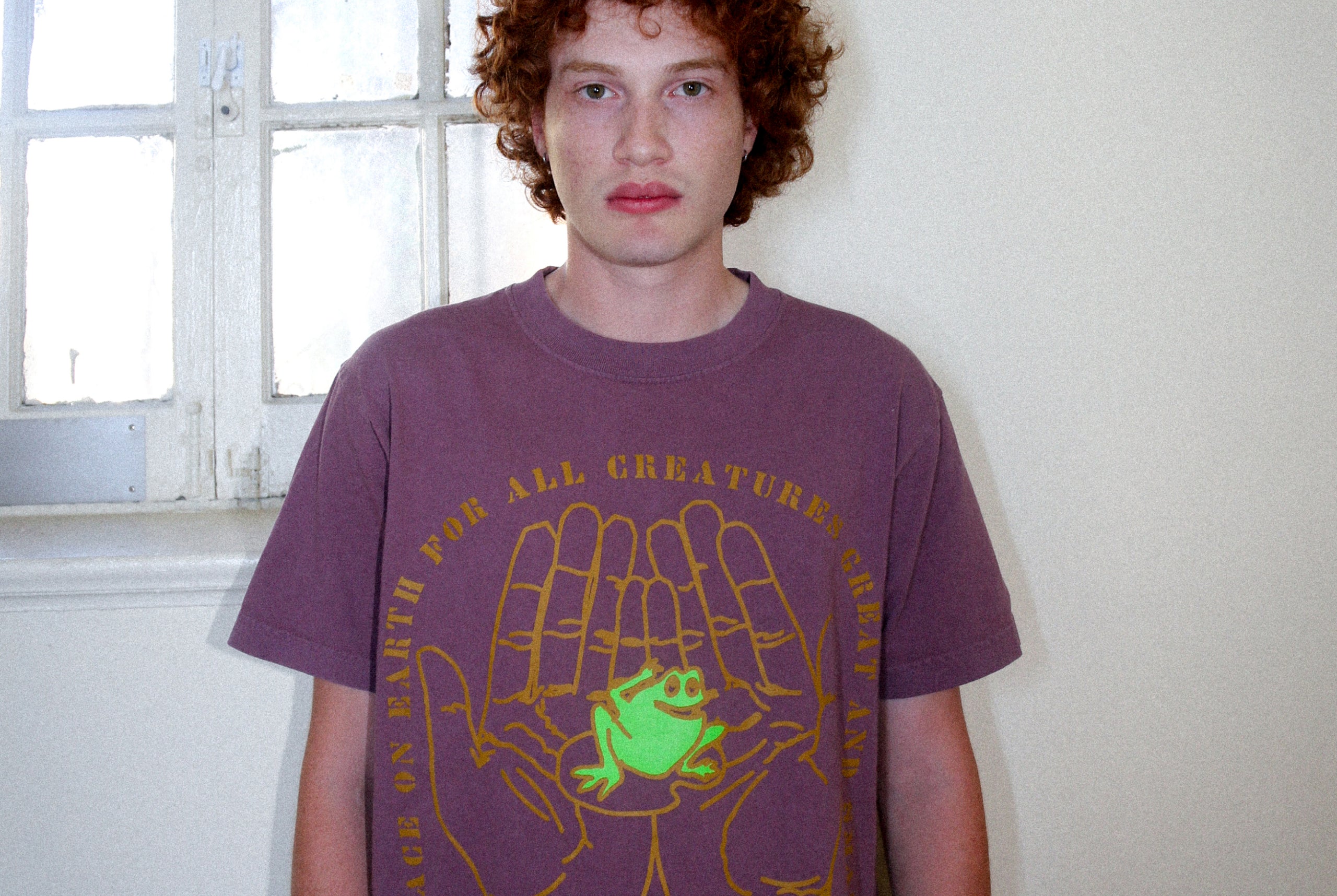 A person with curly red hair wearing a purple graphic t-shirt featuring a green frog and gold handprint design, standing indoors against a light-colored wall and window.