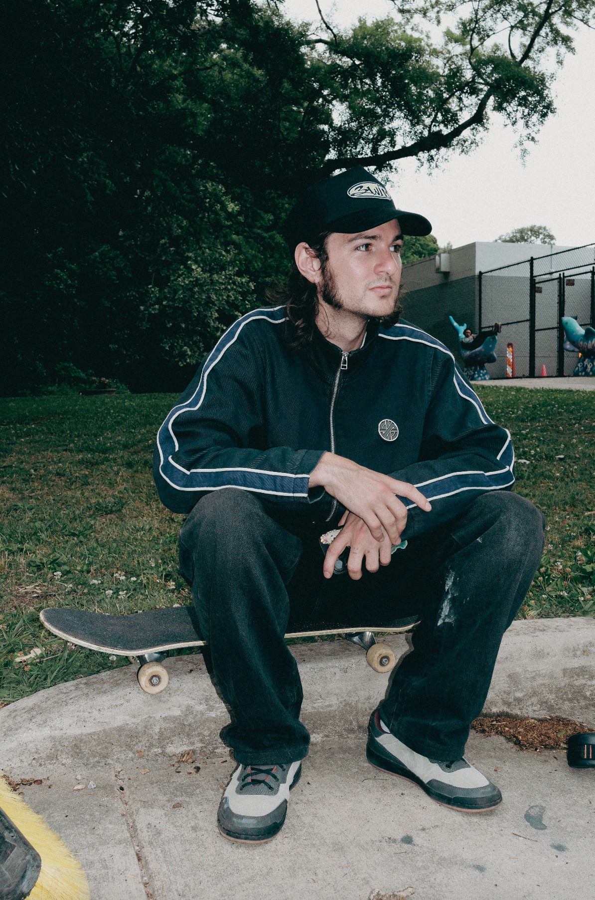 Person sitting on a skateboard wearing a jacket and cap in an outdoor setting.