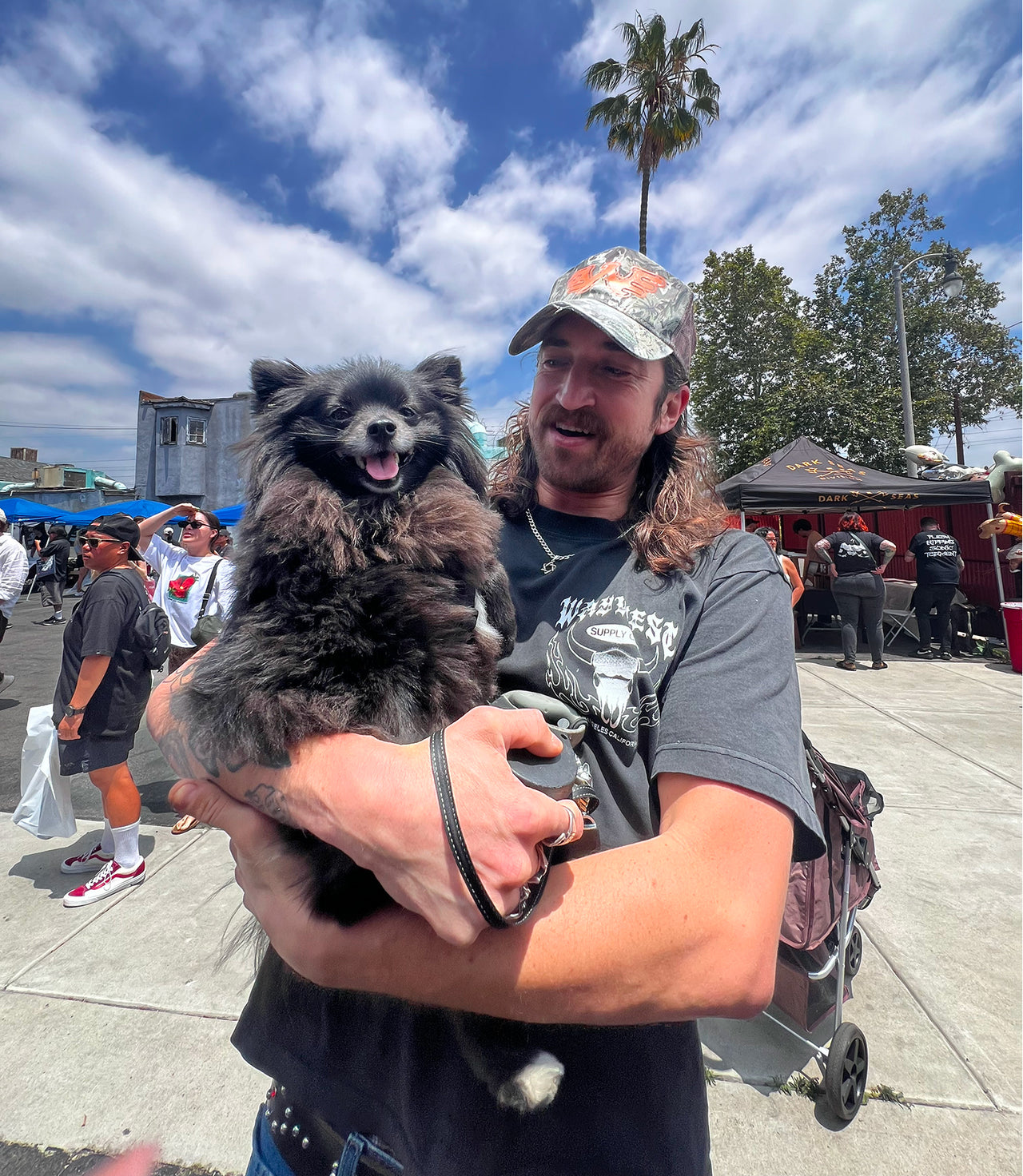 Man holding a small dog outdoors with palm trees and people in the background
