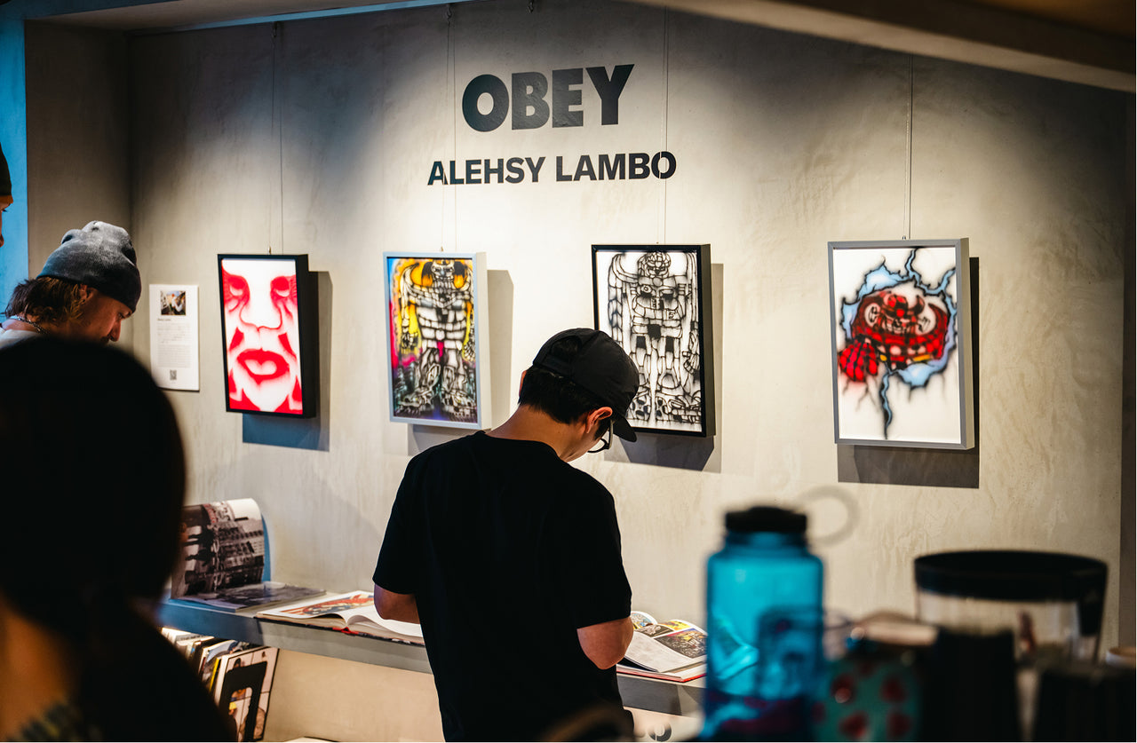 Art exhibition with 'OBEY' and 'Alehsy Lambo' artwork on a wall.
