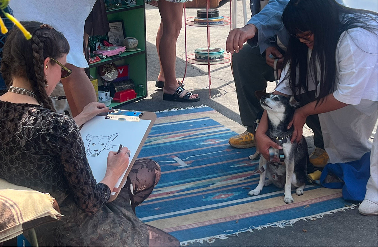 Person drawing a dog on a colorful mat with another person petting a dog nearby.